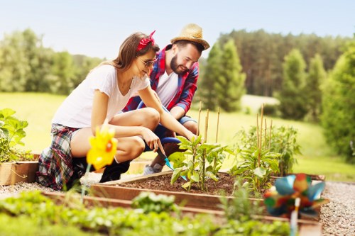 A man and a woman are working together in a well-maintained outdoor garden, tending to a raised flower bed filled with young plants and soil. The woman is using a small gardening tool while the man, wearing a straw hat and a plaid shirt, observes attentively. Surrounding the flower bed are various garden elements, including small decorative plant markers and a colourful toy watering can. The garden features neatly kept grass on the lawn and a backdrop of lush green trees under bright, natural sunlight, indicating clear weather. The scene portrays a peaceful and productive outdoor space likely situated in a residential area near Archway, with a focus on gardening activities, maintenance, and landscape enhancement typical of professional gardening services.