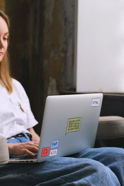 A close-up view of a laptop placed on a person's lap, with various colorful stickers including a yellow machine-readable code, a red power button, and programming language labels such as PHP and HTML.